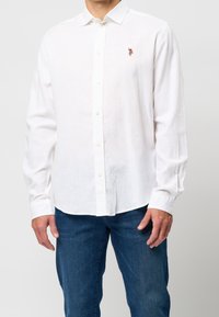 Valkoinen pitkähihaisen pellavapaita, jossa on nappikiinnitys ja pieni kirjailtu yksityiskohta rinnassa, yhdistetty tummansinisiin farkkuihin.