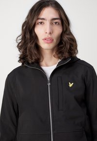 Jachetă neagră cu fermoar și glugă, cu un logo galben brodat pe piept și un design cu buzunar frontal. Textură netedă, material ușor.