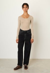 Beige long-sleeve top, loose-fitting black denim jeans with visible stitching, and dark brown laced shoes on a light gray floor.