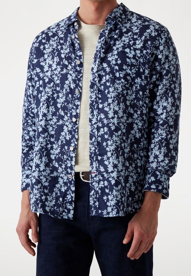 Homme portant une chemise bleu marine à boutons avec un motif floral bleu clair, un t-shirt beige en dessous et un jean foncé.