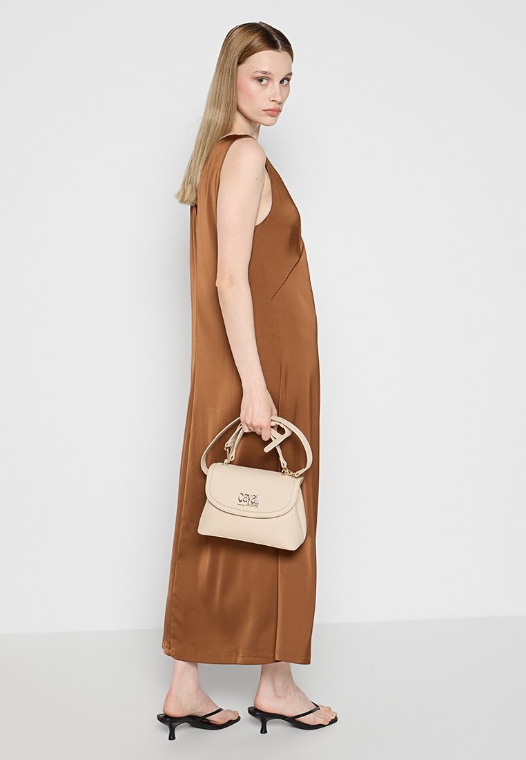 Bruine satijnen mouwloze jurk, vloeiende ontwerpen. Model houdt een crèmekleurige handtas vast met een gouden logo en een gestructureerde vorm. Zwarte hakken sandalen.