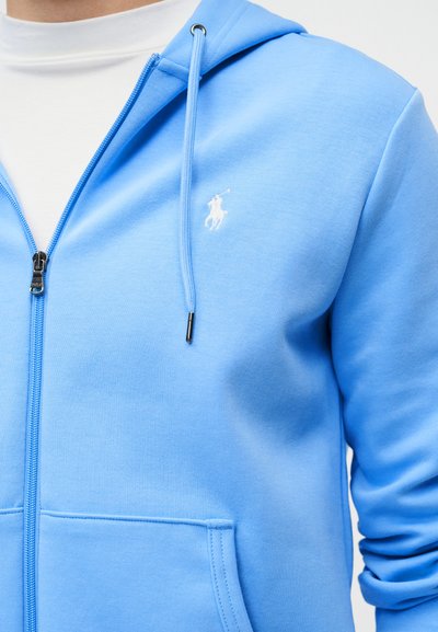 Άνδρας που φοράει ανοιχτό μπλε φούτερ με φερμουάρ και λευκό λογότυπο παίκτη πόλο στο στήθος, πάνω από λευκό μπλουζάκι, με φόντο μονόχρωμο.