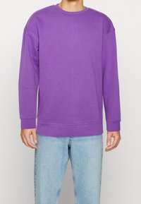 Sudadera morada de manga larga, con cuello redondo y puños de canalé; presenta una textura suave y un corte ligeramente relajado. Combinada con jeans de color azul claro.