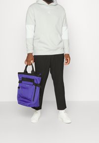Persona que lleva una sudadera ligera gris, pantalones negros, zapatillas blancas, sosteniendo una bolsa tote púrpura y negra con cremalleras y correas laterales.