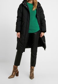 Černá péřová bunda s kapucí a zipem, zelený žebrovaný svetr, černé kožené kalhoty a hnědé kotníkové boty se vzorem hadí kůže.