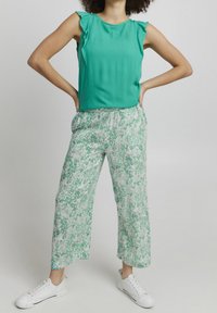 Top verde sin mangas con hombros rizados combinado con pantalones blancos que tienen un patrón floral verde y zapatillas blancas.