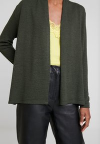 Olivgrüner Cardigan mit weicher Textur, offenem Frontdesign und langen Ärmeln, kombiniert mit einem leuchtend gelben, mit Spitze verzierten Top und schwarzer Hose.