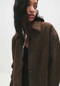 Rjava zamazana srajčna jakna s ovratnikom, ki ima dva temna gumbka in sproščen kroj. Tkanina se zdi mehka in teksturirana.