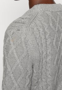 Zbliżenie osoby w jasnoszarym swetrze w warkocz, ukazujące szczegółowe wzory teksturalne na ramieniu i plecach.