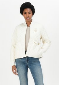 Gepolsterte cremefarbene Bomberjacke mit gerippten Bündchen, Reißverschluss vorne und zwei Seitentaschen. Mit goldfarbenen Beschlägen und einem dezenten Markenlogo.