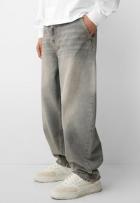Light grey wide-leg jeans made of denim, featuring a faded texture. Paired with white and tan sneakers, showcasing a relaxed fit.