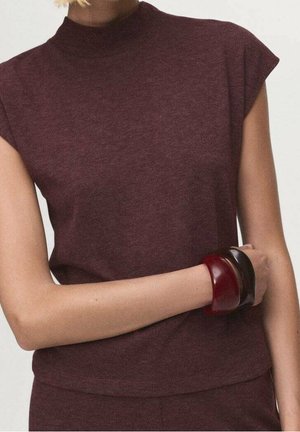 Personne portant un haut en maille sans manches marron foncé avec un col montant et deux bracelets épais, un rouge foncé et un marron foncé, au poignet.