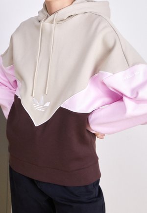 Persoon draagt een colorblock-hoodie met beige, roze en bruine delen en het Adidas-logo op de borst en mouw.