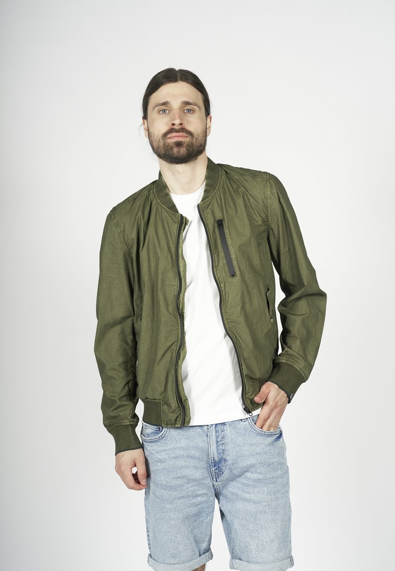 Giacca bomber verde oliva con zip frontale e tasche laterali. Tessuto leggero con polsini a coste, indossata sopra una maglietta bianca e pantaloni corti di jeans.