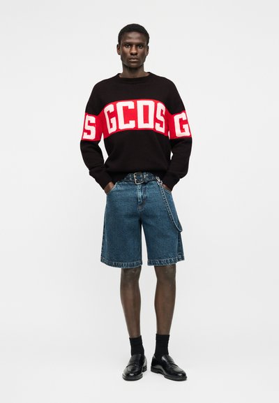 Άνδρας που φοράει μαύρο πουλόβερ με κόκκινα και λευκά γράμματα "GCDS", τζιν σορτς με ζώνη και αλυσίδα, μαύρες κάλτσες και μαύρα δερμάτινα παπούτσια.