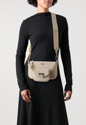 Sac bandoulière beige en cuir texturé, avec une poche avant, des accessoires en métal argenté et une sangle avec le logo.
