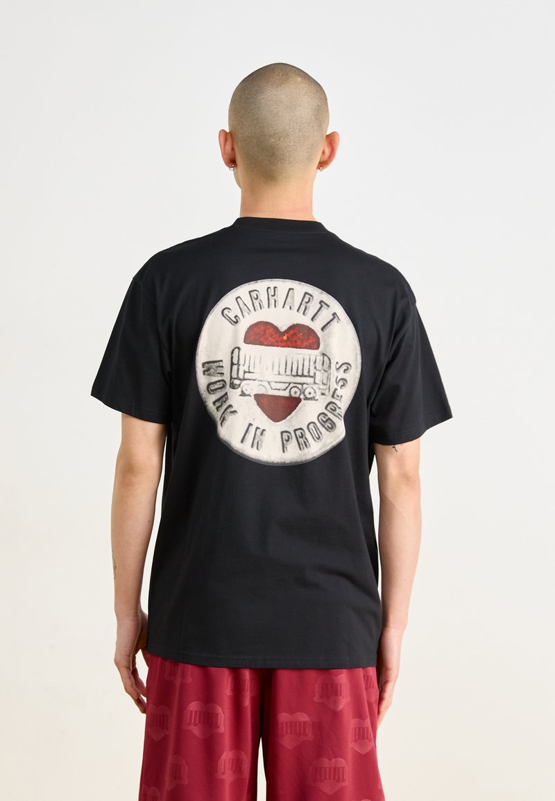 Sort T-shirt med et cirkulært grafisk design på ryggen med "Carhartt" og "Work in Progress" samt et rødt hjerteaccent. Kortærmet, lavet af bomuldsmateriale.