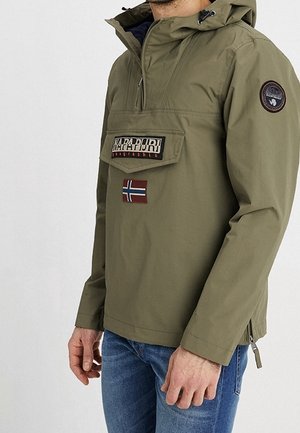 Mężczyzna w oliwkowo zielonej kurtce z kapturem z przodu kieszenią, naszywką norweskiej flagi oraz okrągłą naszywką na lewym rękawie, w połączeniu z niebieskimi dżinsami.