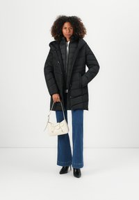 Zwarte pufferjas met een capuchon, voorzien van een gewatteerd ontwerp, voorzakken en een knoopsluiting, gecombineerd met uitlopende blauwe jeans en een witte handtas.