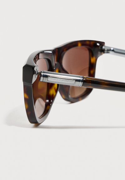 Γυαλιά ηλίου με καφετί φακούς και ασημένιους μεταλλικούς βραχίονες, χαραγμένους με το λογότυπο "Dolce & Gabbana" σε λευκό φόντο.