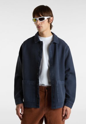 Chaqueta texturizada de color azul marino con cierre de botones, cuello extendido y dos bolsillos frontales. Combinada con una camiseta blanca y pantalones marrones.