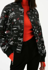 Giacca bomber nera con grafiche bianche e rosse, cuori e testo. Polsini e orlo a coste. Indossata sopra un dolcevita rosso a coste.