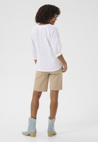 Witte linnen blouse met geplooide halslijn, beige shorts en lichtblauwe cowboylaarzen met decoratieve stitching. De outfit heeft een ontspannen pasvorm.