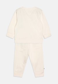 Tuta color crema realizzata in morbido tessuto. Presenta maniche lunghe, polsini a costine e una tasca posteriore sui pantaloni.