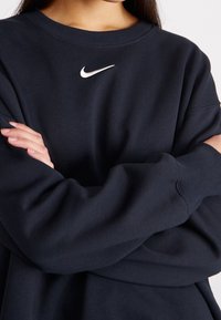 Navy blue sweatshirt with a round neckline and long sleeves. Features a white Nike logo on the chest and ribbed cuffs. Smooth texture.