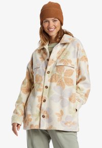Fleece shirt jacket with oversized fit, cream background, large floral patterns in orange, green, and purple, and wooden buttons.