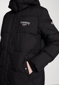 Černá péřová bunda s logem "tommy jeans" na hrudi, malým vlajkovým emblémem na rukávu a rukou v postranní kapse na cvoky.