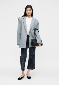 Femme portant un blazer gris clair oversized, un haut blanc, un pantalon court à motifs, des escarpins slingback noirs, tenant un sac à main noir sur fond blanc.