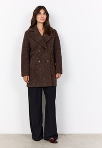 Brauner doppelt geknöpfter Mantel aus strukturiertem Stoff, mit Reverskragen und vorderen Knöpfen, kombiniert mit schwarzen weit geschnittenen Hosen.