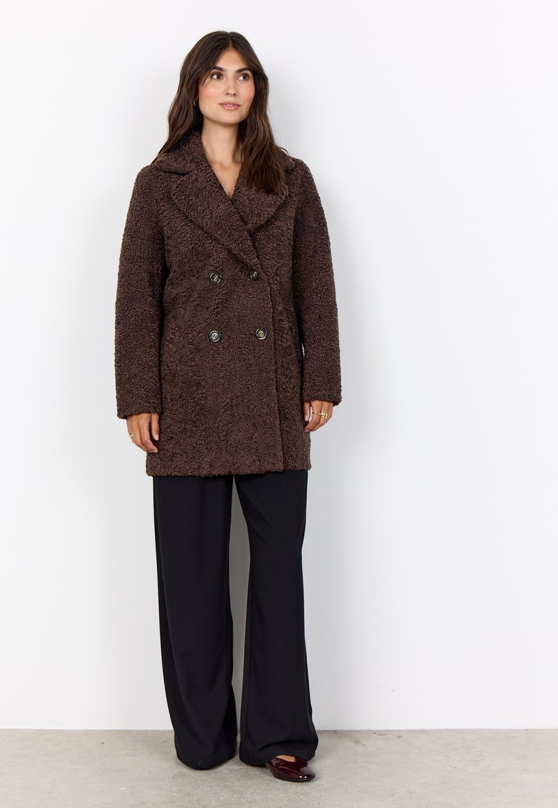 Brauner doppelt geknöpfter Mantel aus strukturiertem Stoff, mit Reverskragen und vorderen Knöpfen, kombiniert mit schwarzen weit geschnittenen Hosen.