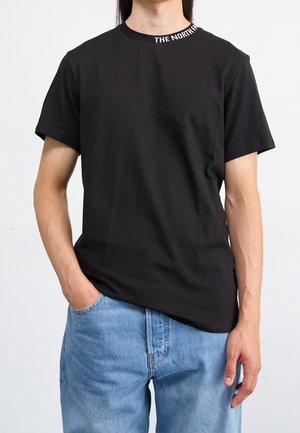 Person iført en sort t-shirt med korte ærmer med teksten "THE NORTH FACE" på kraven og lyseblå denimjeans, den ene hånd i lommen.