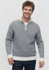 Leende man som bär en grå och vit mönstrad tröja med kvartslång dragkedja och mörka jeans, står mot en plain ljus bakgrund.