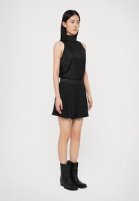 Vrouw die staat in een mouwloze zwarte top met hoge hals, zwarte korte broek met riem en zwarte laarzen tot halverwege de kuit tegen een witte achtergrond.