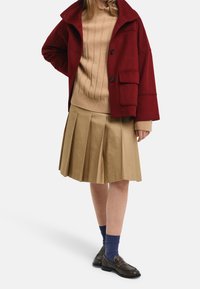 Donna che indossa una giacca rossa con bottoni, un maglione a coste beige, una gonna plissettata beige, calzini blu e mocassini marroni scuri, in piedi con una mano sulla vita.