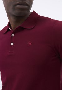 Bordeaux poloåben med en struktureret stof, tre knapper og et lille broderet fuglelogo på venstre bryst. Klassisk kravedesign.