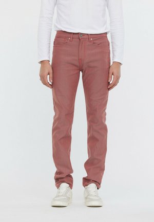 Jeans à motifs rouges avec une coupe slim, dotés de coutures orange et d'un bouton de fermeture. Portés avec une chemise blanche à manches longues et des baskets blanches.