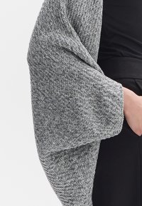 Dzianinowy szal w jodełkowy wzór, utrzymany w odcieniach szarości i miękkiej teksturze, zarzucony na czarną odzież, z szerokimi, luźno opadającymi rękawami.