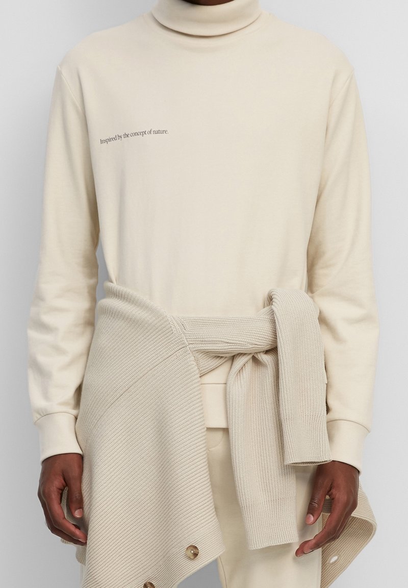 Pull à col roulé beige avec le texte « Inspiré par le concept de la nature. », associé à un pull en maille beige noué à la taille.