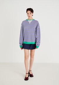 Maglione oversize a righe in viola e bianco, con orlo a costine verde. Abbinato a una mini gonna di jeans decorata con ricami rossi.