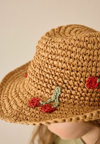 Chapeau tissé en paille de couleur beige, avec un bord structuré, arborant des broderies florales rouges et des accents verts. Surface texturée avec une finition naturelle.