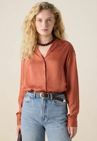 Rust-colored silk shirt with a collar, long sleeves, and buttons, paired with light blue denim jeans and a brown belt. Dark beaded necklace included.