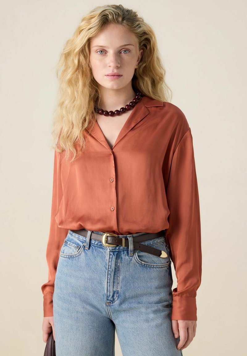 Rust-colored silk shirt with a collar, long sleeves, and buttons, paired with light blue denim jeans and a brown belt. Dark beaded necklace included.