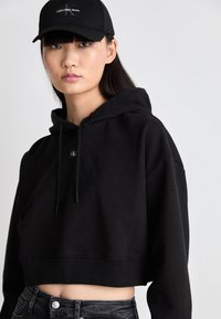 Black cropped hoodie with a drawstring hood, ribbed hem, and subtle logo detail. Worn with black high-waisted jeans.