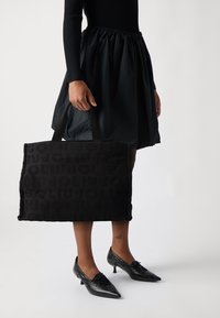 Borsa tote in tessuto nero con design del logo in rilievo, forma rettangolare e manici corti neri, tenuta accanto a una gonna nera a ruota e scarpe con punta.