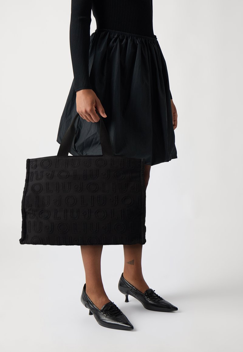Borsa tote in tessuto nero con design del logo in rilievo, forma rettangolare e manici corti neri, tenuta accanto a una gonna nera a ruota e scarpe con punta.