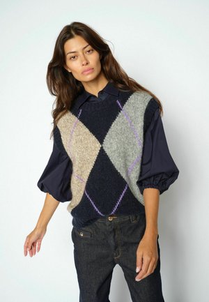 Woman with long brown hair wearing a navy shirt with puffed sleeves and a sleeveless argyle sweater vest in navy, beige, and gray.